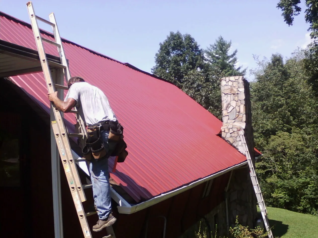 Roofer on a ladder beside a red metal roof and stone chimney — Soffit & Fascia Repair by Roofing Brothers in Perry Hall